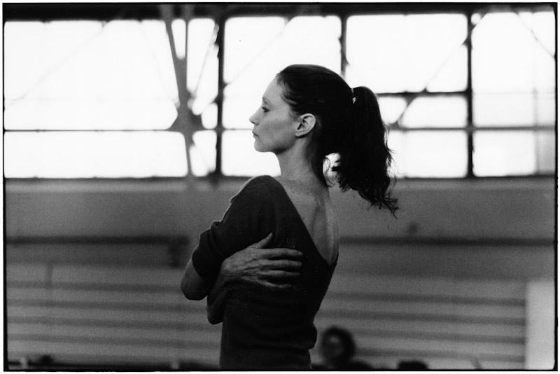 A ballerina with a ponytail stands in profile, arms folded across herself, against a backdrop of large windows