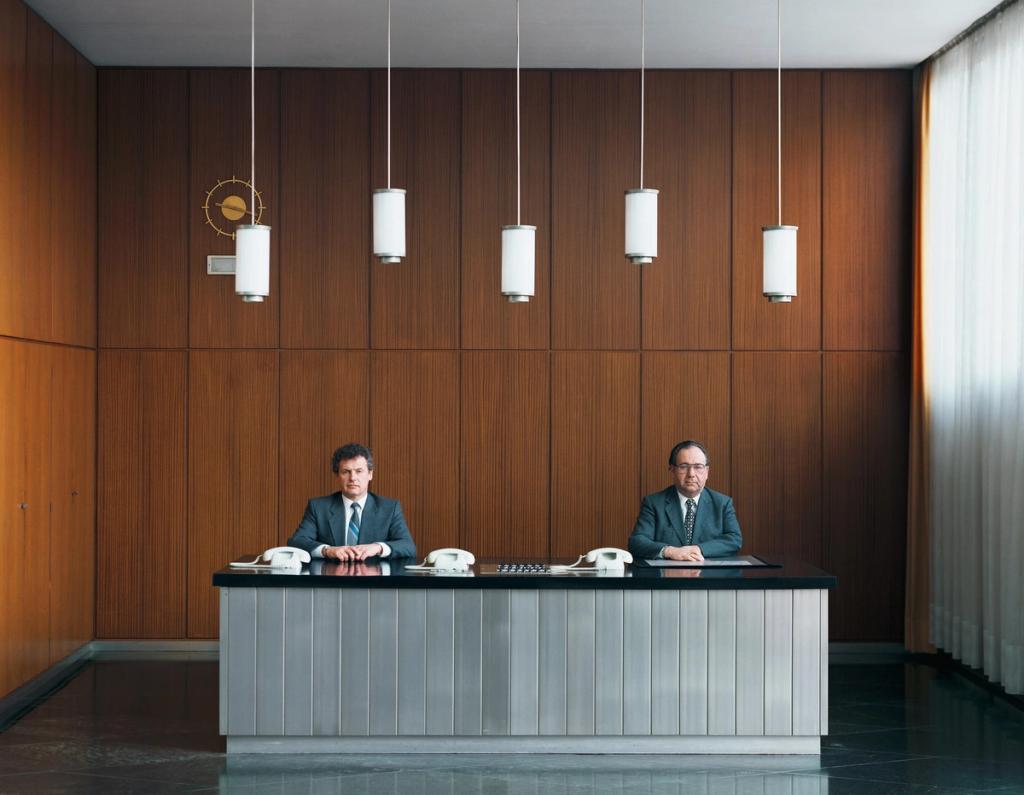 Two men in suits at a reception desk in a wood-panelled lobby with pendant lights
