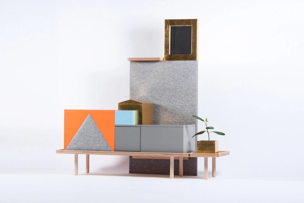 A multi-tiered chest-of-drawers installation in grey felt with orange and brass blocks, set on a wooden bench