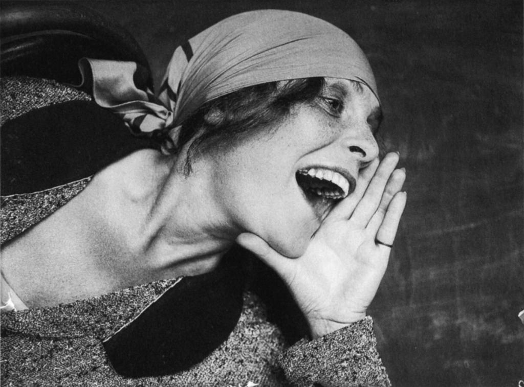 A black-and-white photograph of a woman in a headscarf, screaming, her palm raised to her mouth