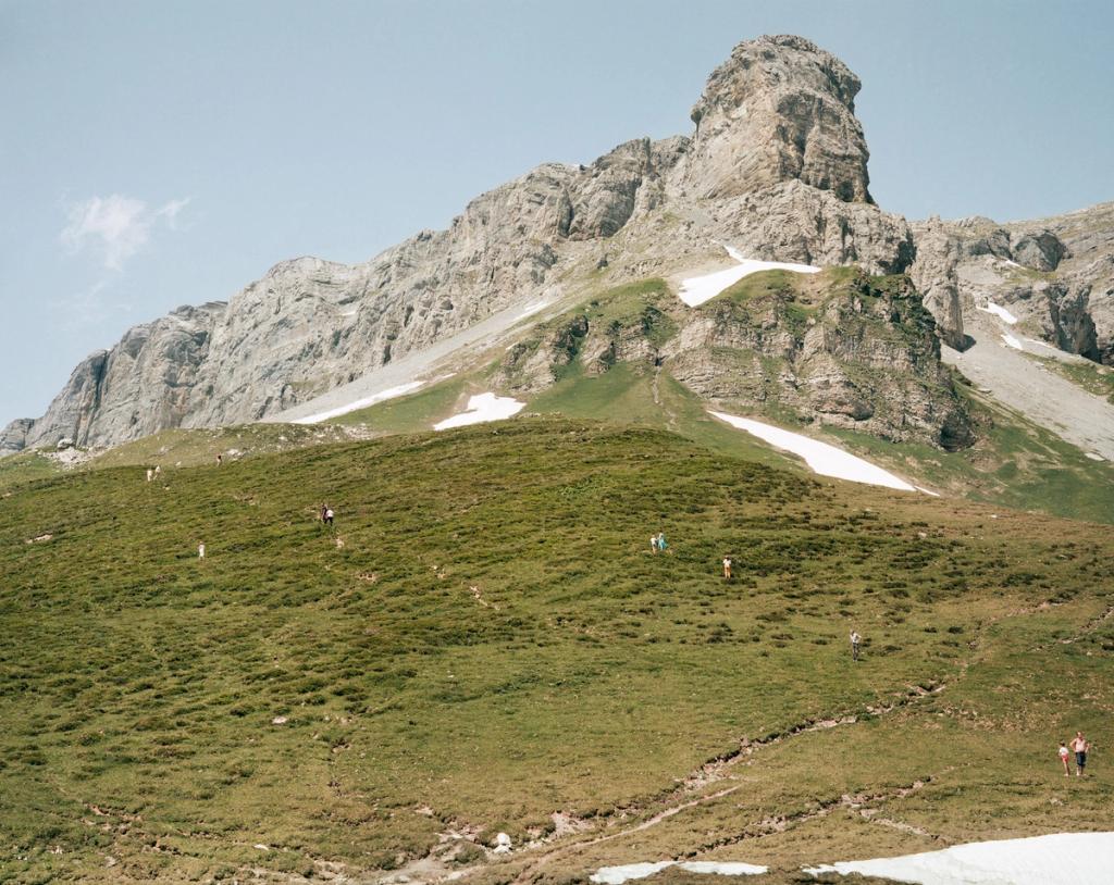 A rocky mountain summit and green alpine slope with tiny figures of hikers