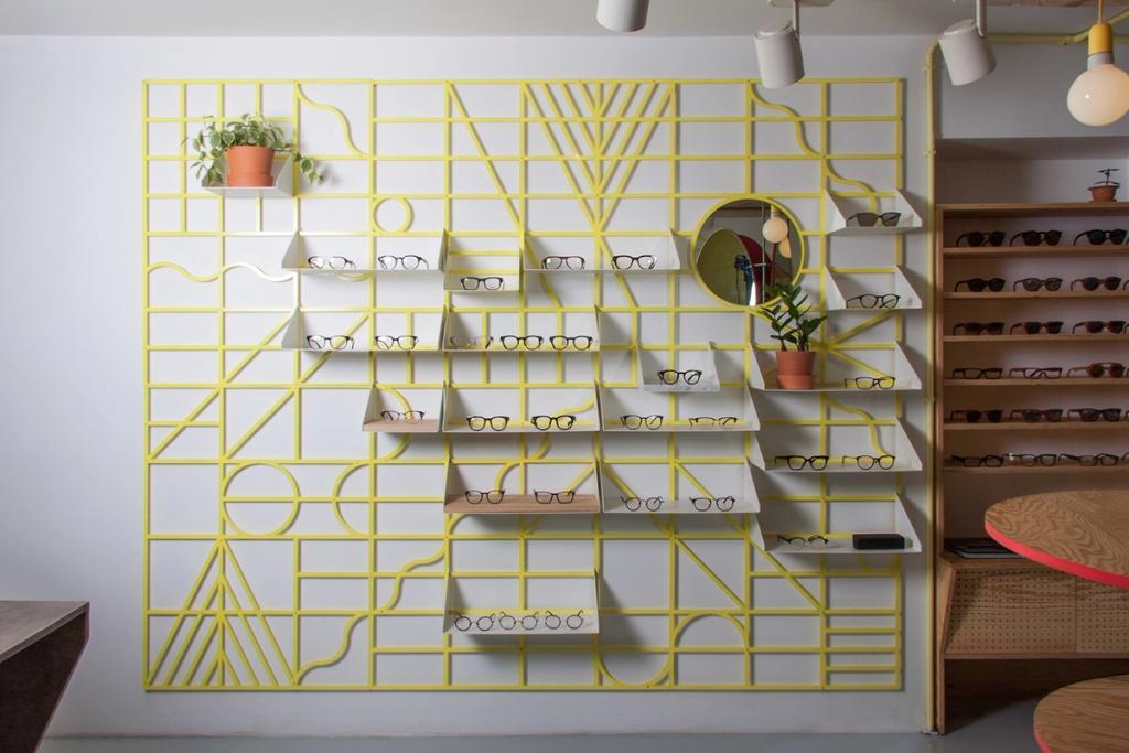A yellow open-work shelving unit on the wall of an optician's, with glasses displayed and a round mirror