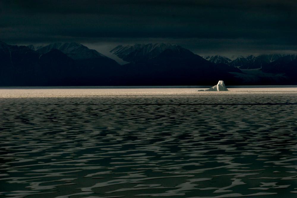 A solitary iceberg on a strip of lit water amid dark mountains and a overcast sky