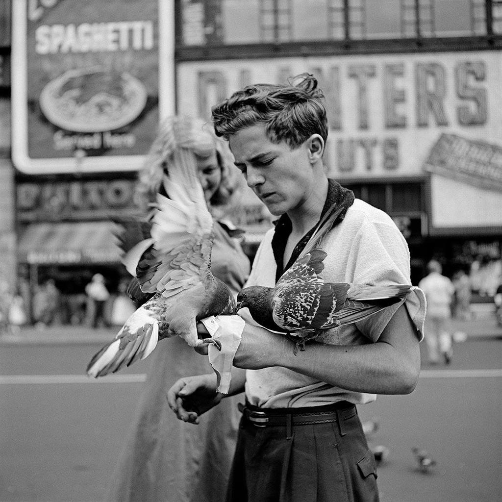 Black-and-white photograph: a young man holds two pigeons with outstretched wings in a public square