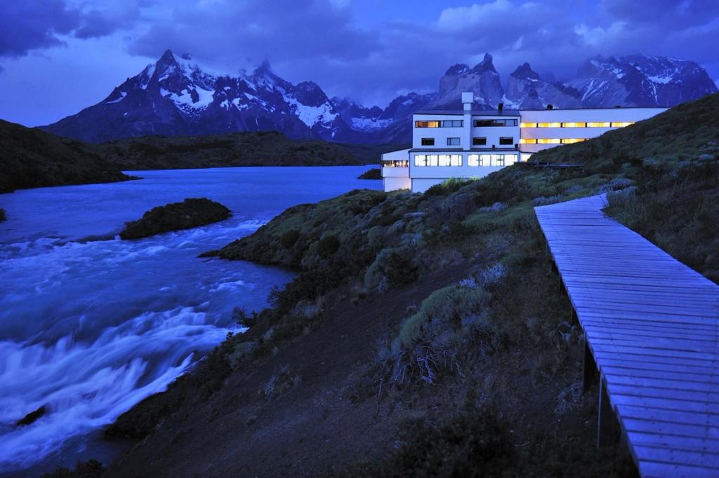 The illuminated Explora Patagonia building at dusk, beside a mountain river and the Paine peaks