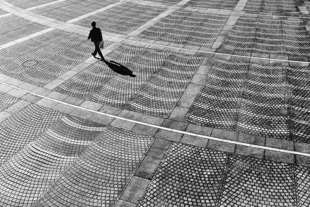 A solitary figure with a long shadow crosses a cobblestoned square, seen from above