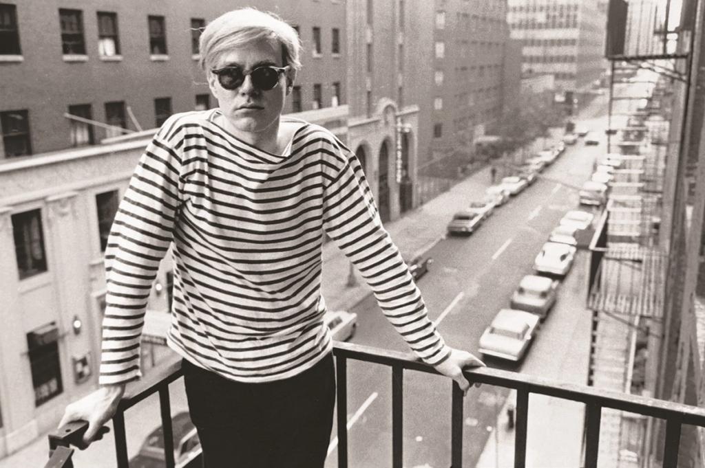Black-and-white photograph of Andy Warhol in a striped sweater and dark glasses on a balcony above a New York street