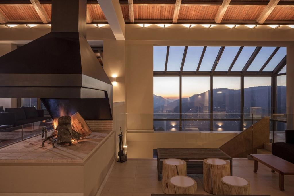 Lounge area with a fireplace and a panoramic window overlooking the mountains of Metsovo at sunset