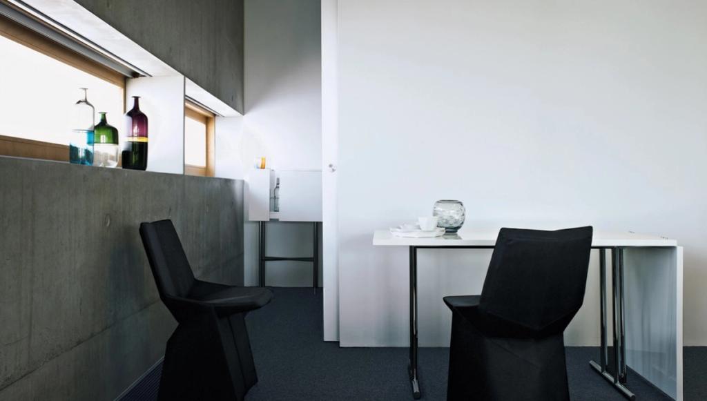 A minimalist dining area with a white table and two chairs in black covers against a concrete wall