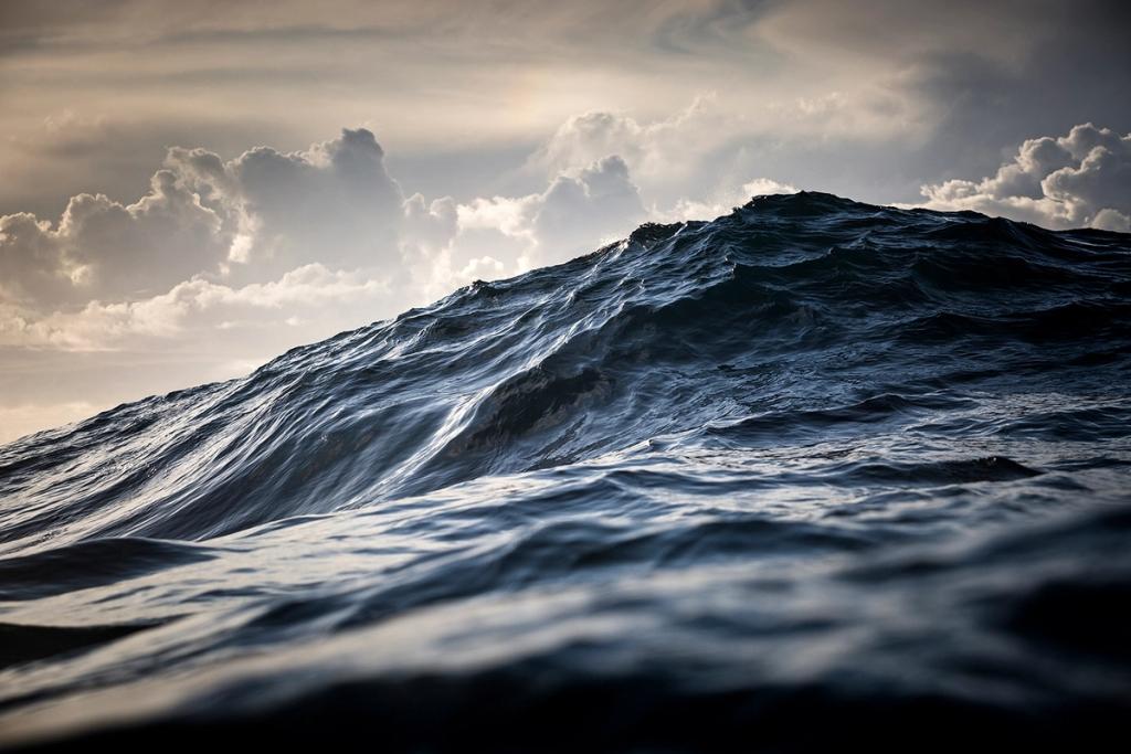 A dark-blue swell on the open ocean beneath clouds pierced by breaking sunlight