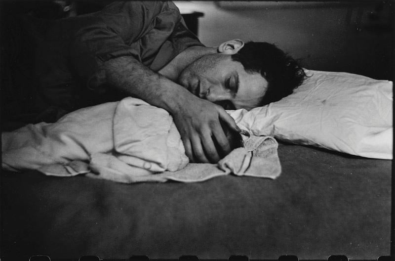 A man sleeps on a bed, arms wrapped around a pillow and a crumpled towel