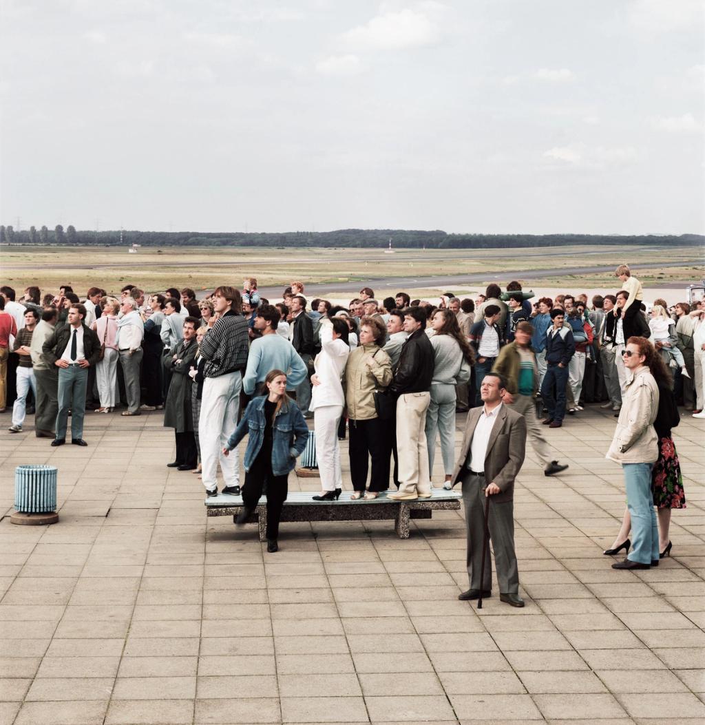 A crowd of people on a concrete esplanade looking upward against the backdrop of a vast open airfield