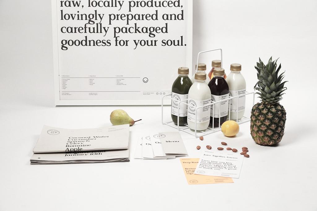 Бутылки соков Leeds Juicery в переноске рядом с ананасом, грушей и плакатом