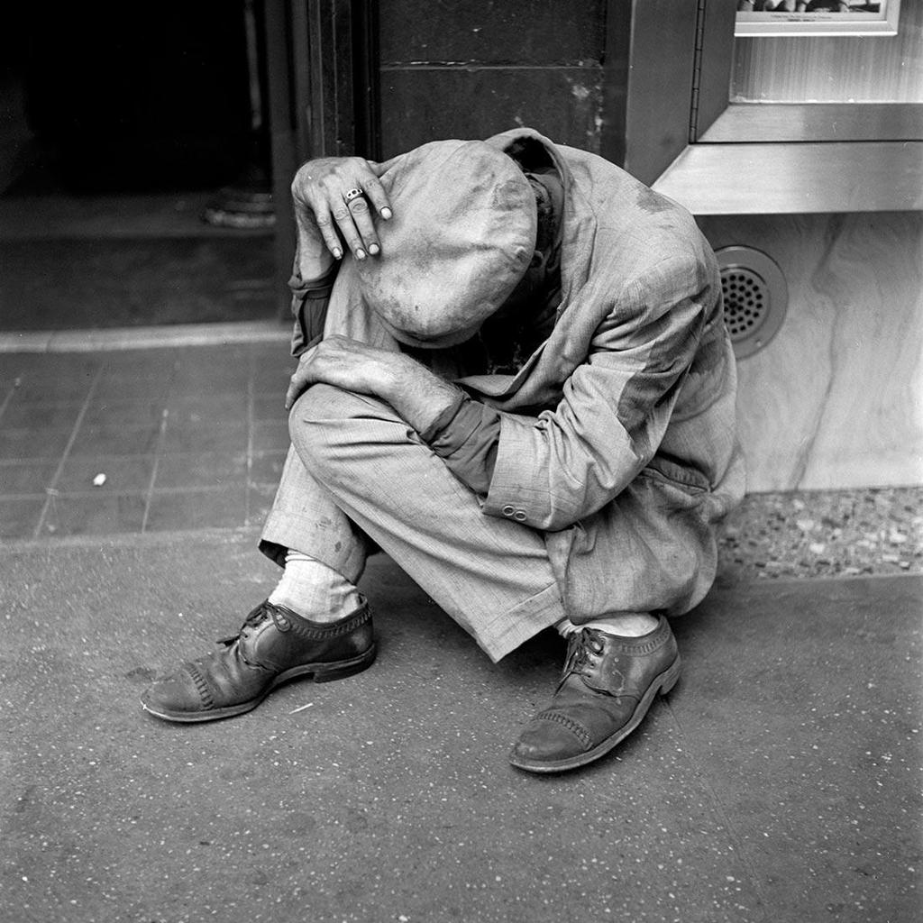 Black-and-white street photograph: a man in a crumpled suit sits on the pavement with his face buried in his knees