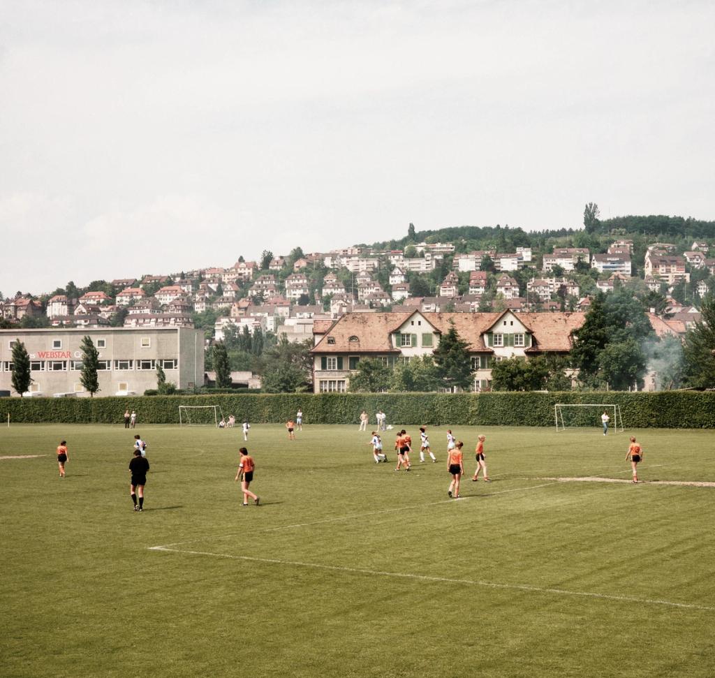 A football match on a green pitch, with dense residential buildings covering the hillside behind it