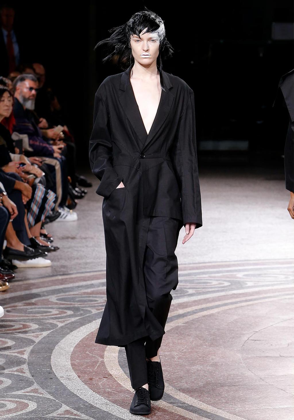 A fitted black jacket and wide trousers with white face paint on the runway