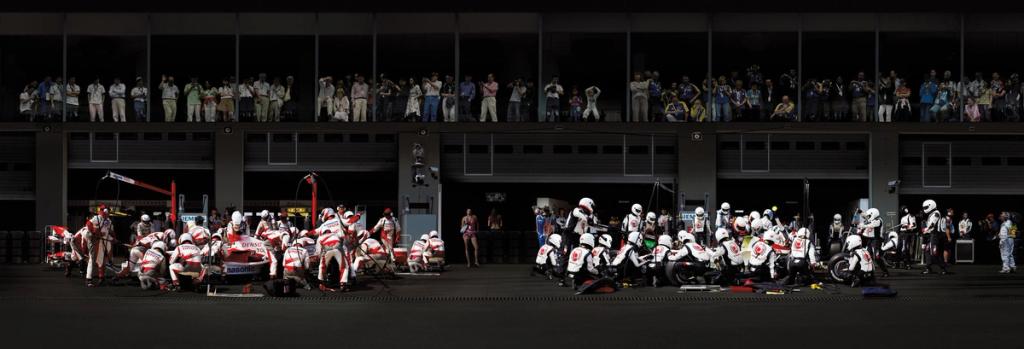 Formula 1 pit-stop panorama: mechanics beside the cars and spectators in the stands, the pit lane half in shadow