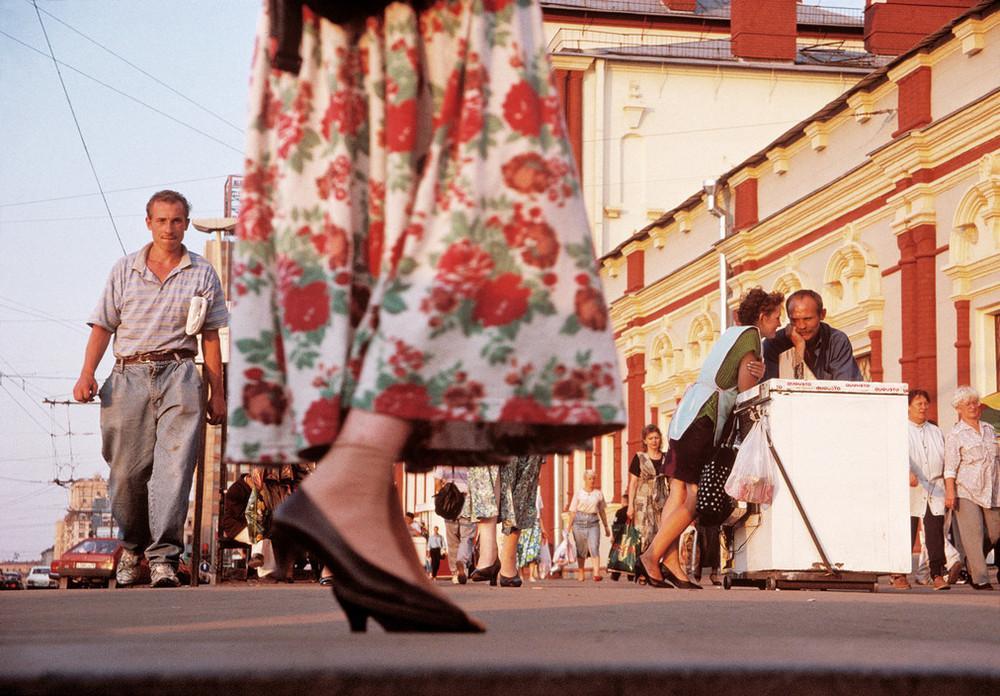 From below: a woman's legs in a floral skirt on a busy summer street lined with vendors