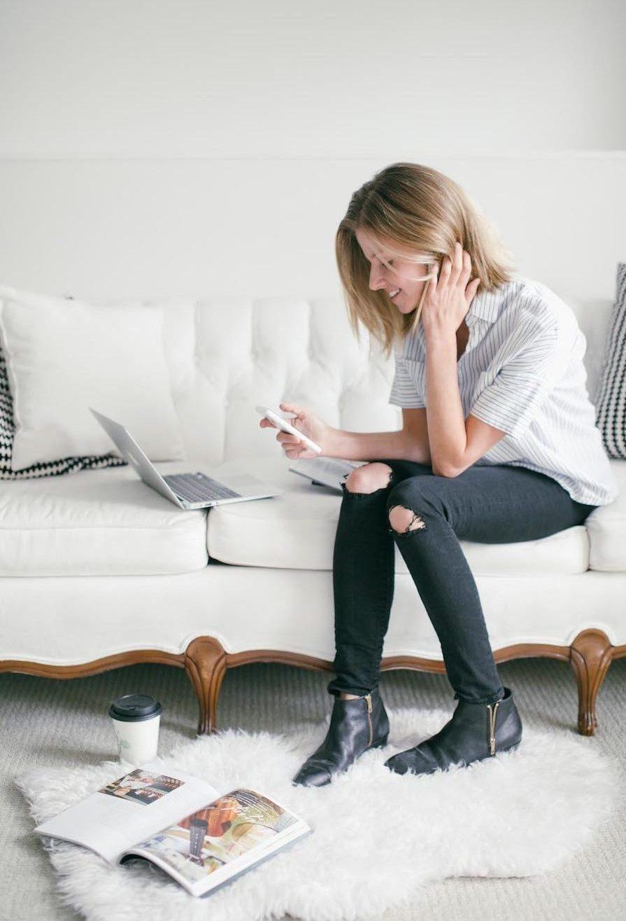 A girl sits on a white sofa with a phone; a laptop and a cup of coffee are nearby