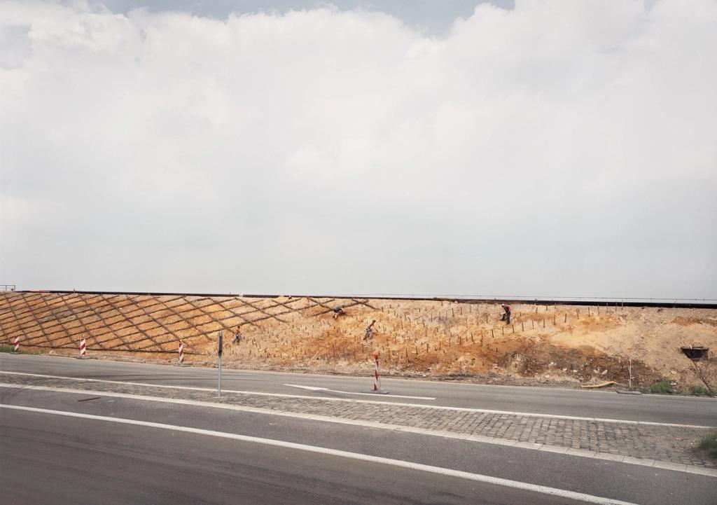A road running along an embankment slope with yellow soil held in place by netting, under a cloudy sky