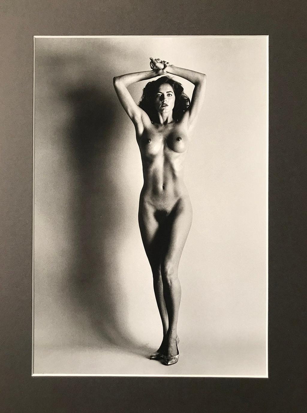 A nude woman in heeled shoes stands with her hands clasped behind her head against a grey wall