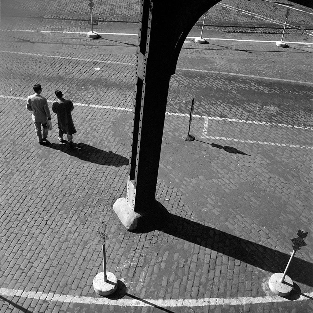 Black-and-white photograph taken from above: two passersby and long shadows on a cobbled square beside an elevated structure's support column