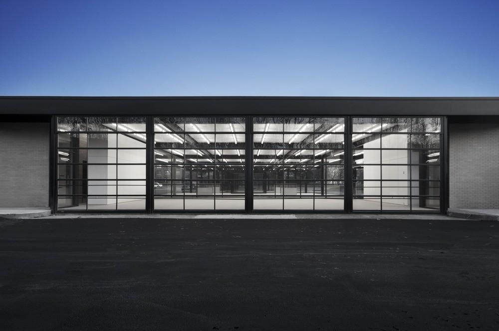 A single-storey glass-and-steel pavilion with a black frame, illuminated at dusk