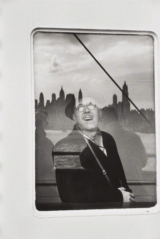 A smiling man in a hat on a ferry, with the silhouette of New York's skyscrapers behind him