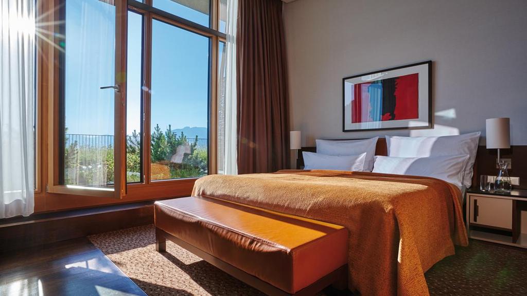 A Kempinski guest room with a bed, leather bench and a window overlooking sun-drenched mountains