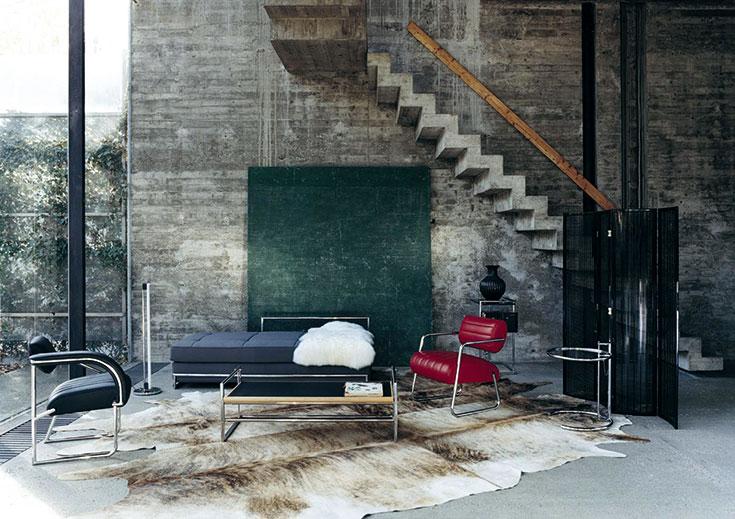 Interior with concrete walls and a staircase: a daybed, a red armchair, a screen and an E-1027 side table on a sheepskin