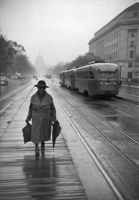 A woman carrying umbrellas walks across a wet cobbled street; the Capitol building and a tram are visible in the distance, in Washington
