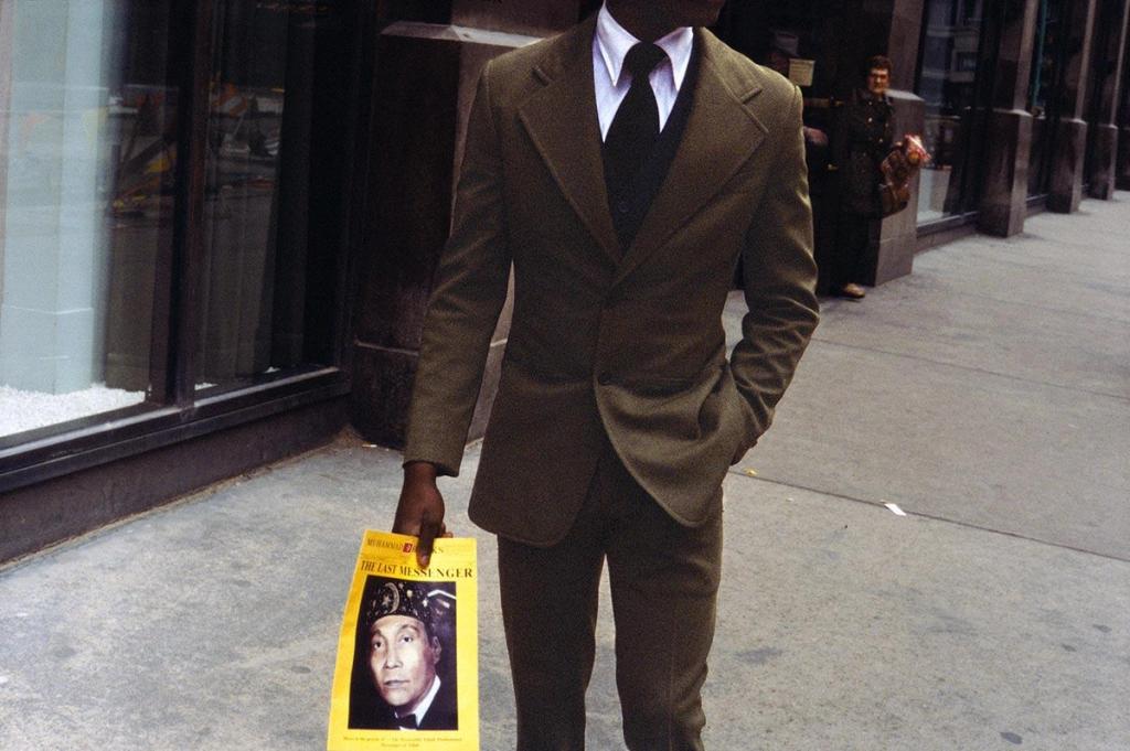 Colour photograph: a man in a green suit walking along the street with a paper bag and a newspaper in his hand