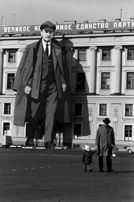 A vast portrait of Lenin on the façade of a building; below it, a man with a child stands on an empty square
