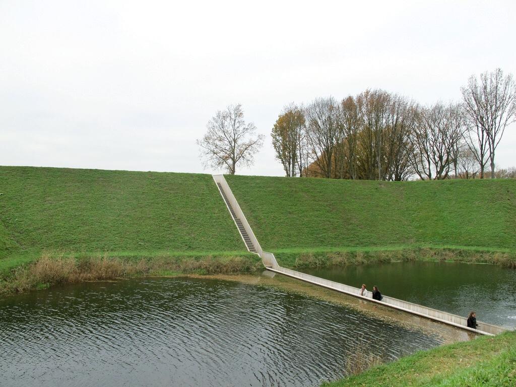 The Moses Bridge from a distance: a narrow timber walkway set within the moat beside a green earthen rampart
