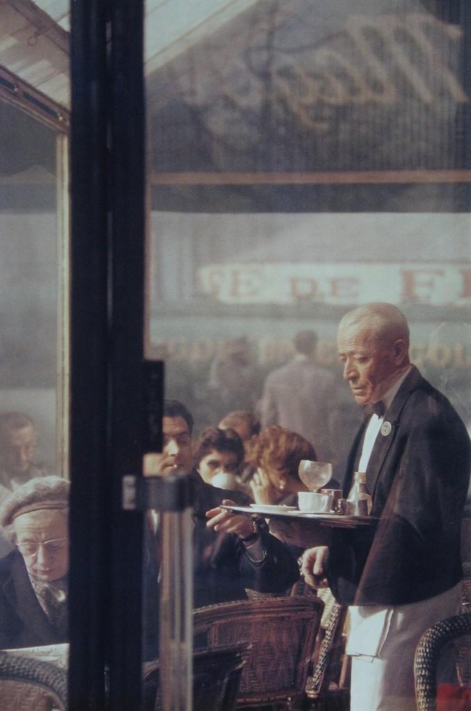 A colour photograph by Saul Leiter: an elderly waiter carrying a tray, seen through the glass of a Parisian café
