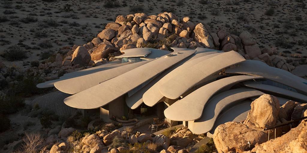 A house with an undulating concrete roof set among desert boulders at sunset, aerial view