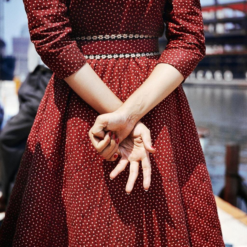 Colour photograph: a woman in a burgundy polka-dot dress stands with her hands behind her back, by a waterfront