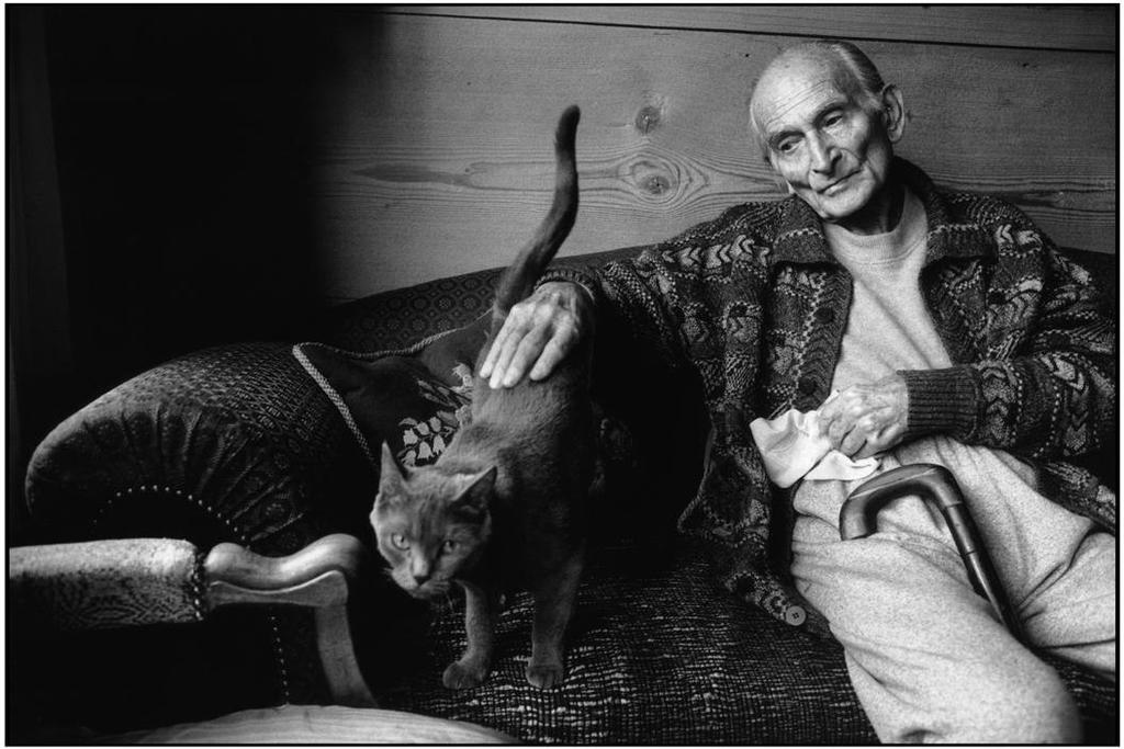 A thin elderly man on a sofa strokes a cat, a walking stick lying beside him