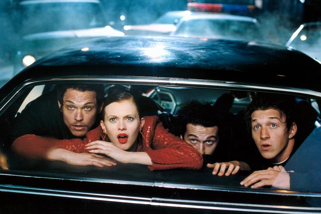Four people in red and dark clothing peer out of the rear window of a car at night