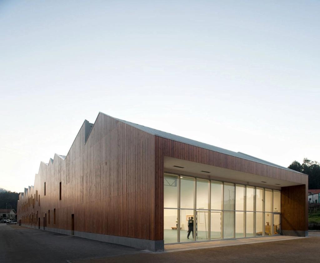 The corner of Mouriz school: timber cladding, a sawtooth roof and a glazed entrance at dusk