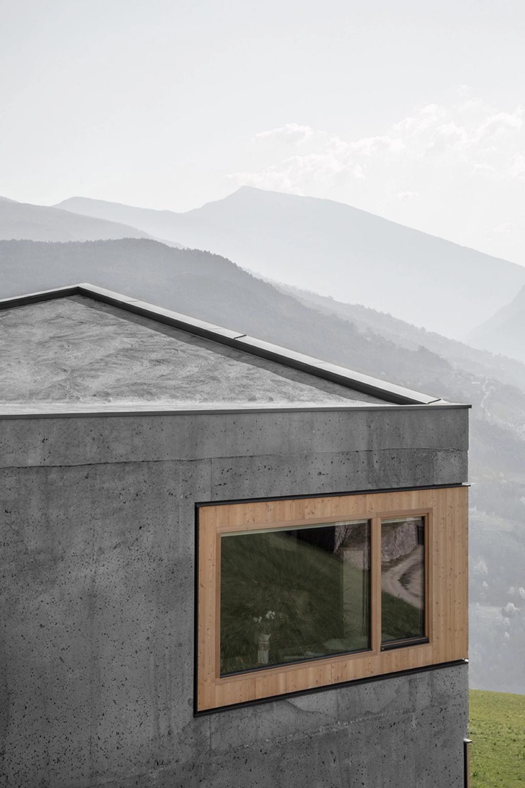 The corner of a concrete house with a wooden window against a haze-shrouded Alpine backdrop