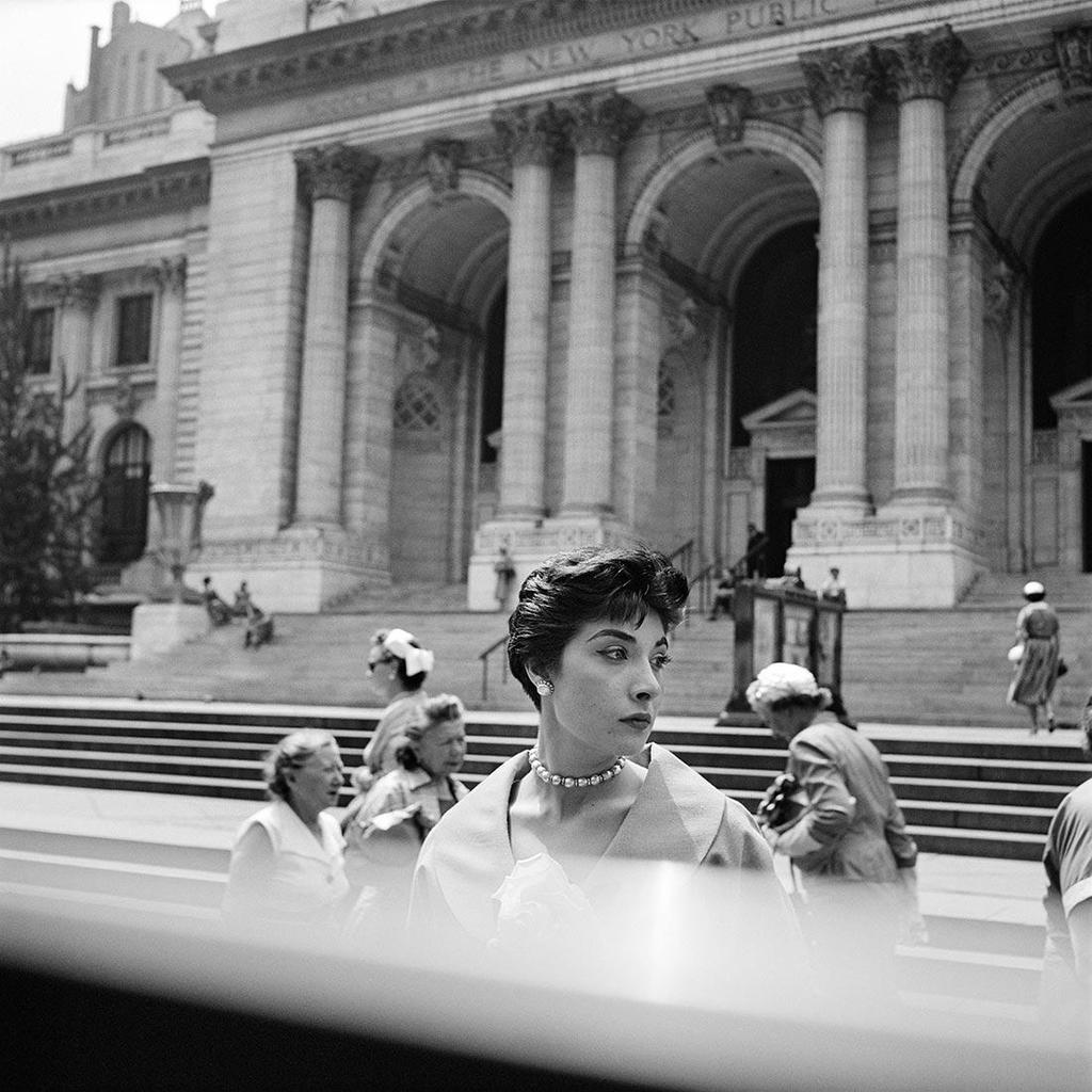 Black-and-white photograph: a woman in a pearl necklace against the colonnade of the New York Public Library