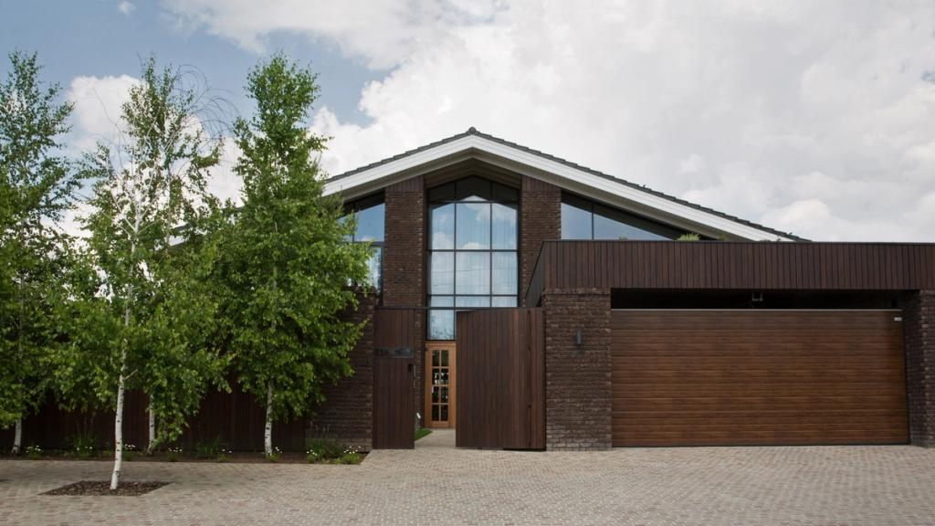 The facade of a dark-brick house with a pitched roof, a glazed gable and wooden garage doors