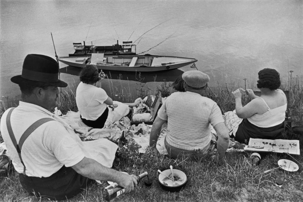 A group of people picnicking by the water, a boat moored on the bank in front of them