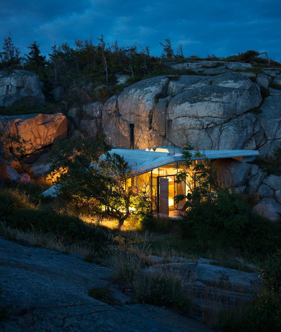 A cabin glowing from within among coastal rocks and trees in the blue dusk