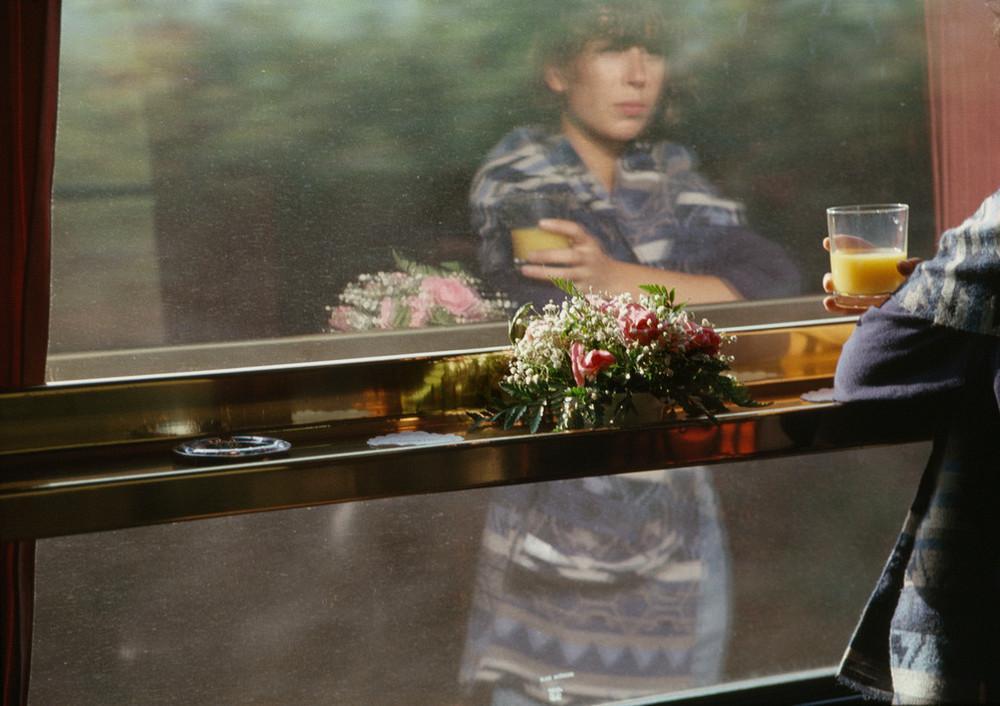 The reflection of a woman holding a bouquet and a glass of juice in a train window