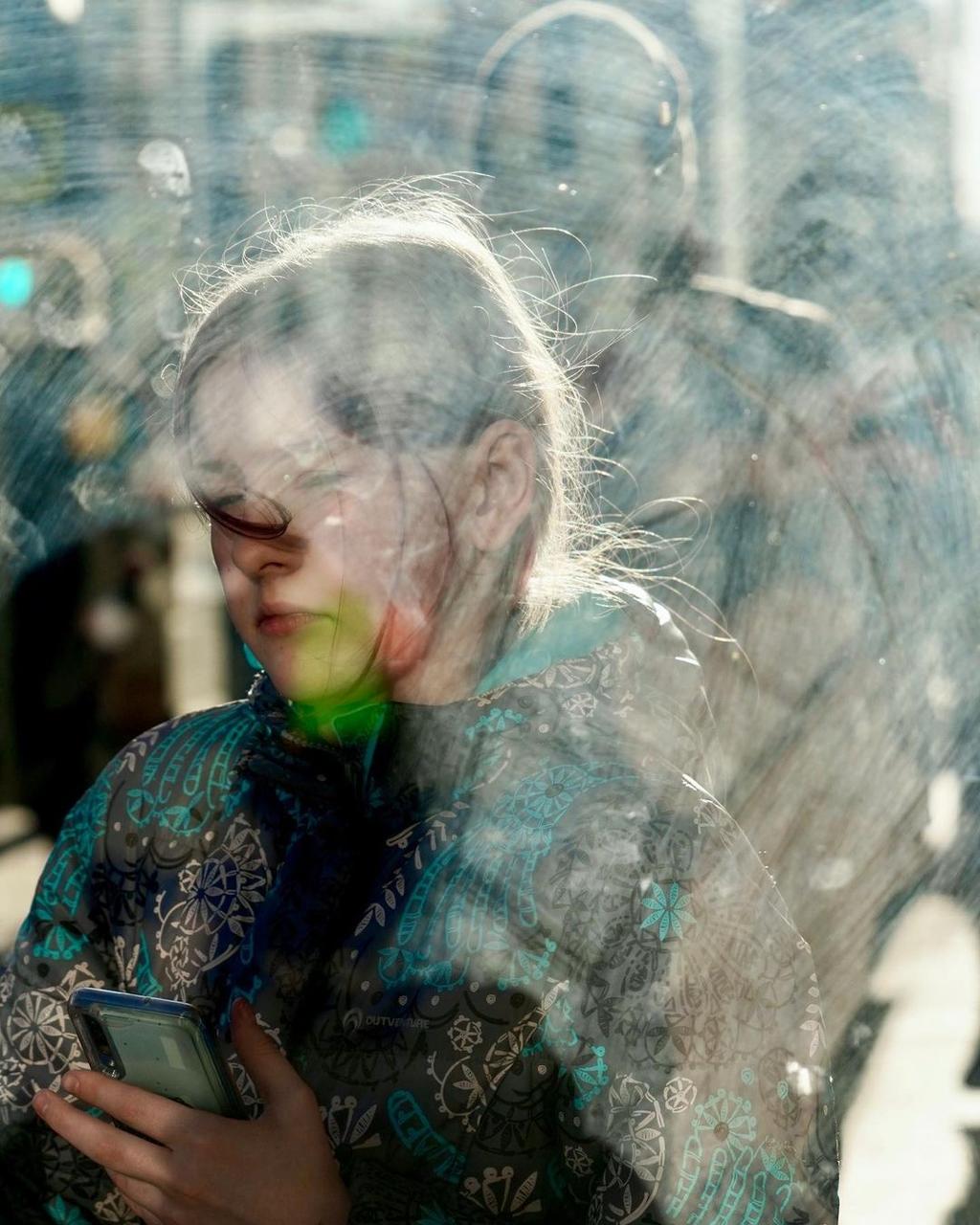 A girl with a smartphone behind glare-streaked glass, her hair streaming in the sunlight
