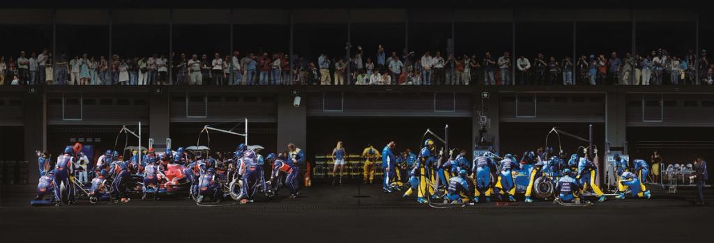 Formula 1 pit-stop panorama: two teams of mechanics in blue-and-yellow overalls servicing the cars