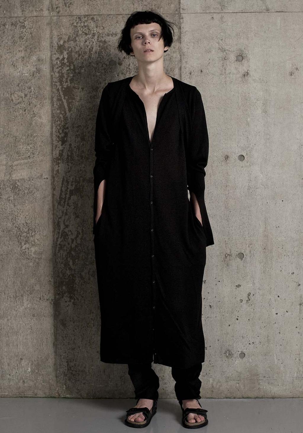 A model by a concrete wall in a long black knit cardigan and sandals