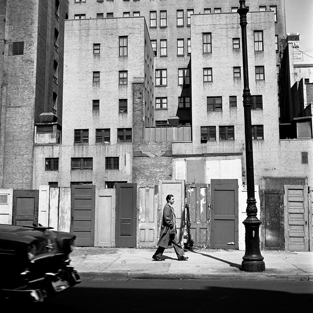 Black-and-white photograph: a solitary man in a coat walks past the weathered doors of a tall building